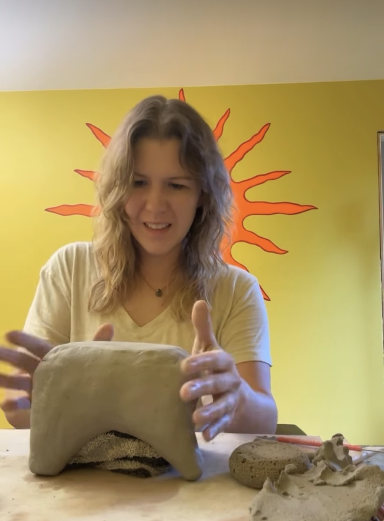 Alisha, the ceramic artist, hand-building an animal form out of raw grey clay in her studio, in front of a sunny yellow wall painted with a large orange folk-art sun.