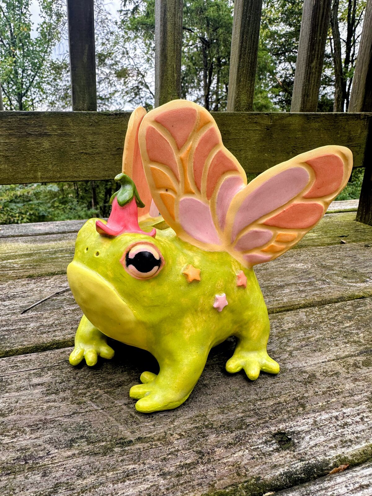 A chartreuse ceramic frog with big pink-rimmed eyes, a pink bellflower cap, pastel star spots, and two pairs of pink, peach, and cream butterfly wings, sitting on a weathered wooden deck.